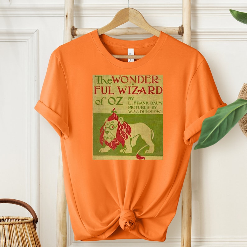 Short sleeve orange shirt featuring vintage book cover of The Wizard of Oz in pale gold with grass green and red font & the cowardly lion wearing glasses. The lion's mane is red and his glasses are green. The lion is set on a light green background.