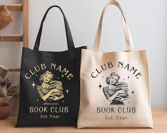 Puede incluir: Dos bolsas de tela con asas negras y blancas. La bolsa negra tiene un diseño dorado de una mujer sosteniendo una pila de libros con el texto "CLUB NAME BOOK CLUB Est. Year". La bolsa blanca tiene el mismo diseño en dorado.