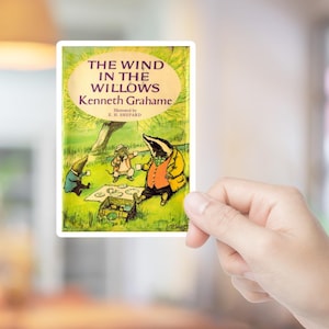 May include: A sticker featuring the cover of "The Wind in the Willows" by Kenneth Grahame, illustrated by E.H. Shepard. The image shows a badger in a yellow coat, a mouse, and a mole, with the title in a gold oval against a green background.