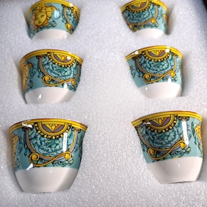 May include: Six small, decorative espresso cups. Each cup features a light blue background with a gold and teal floral design and a white base. The cups are arranged in a white foam insert.