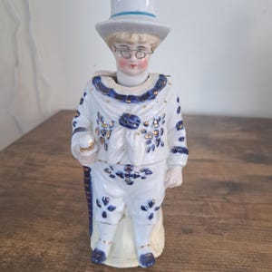 以下が含まれることがあります： 青い帯の白いシルクハット、ワイヤーフレームの眼鏡、青い花柄と金色のアクセントが入った白いセーラー服を着た人物の磁器製フィギュア。右手に小さな物を持っています。