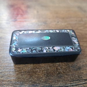 Op de afbeelding: Een klein zwart doosje met een decoratieve rand van iriserende schelpstukken. Het doosje heeft een groene ovale inleg in het midden van het deksel.