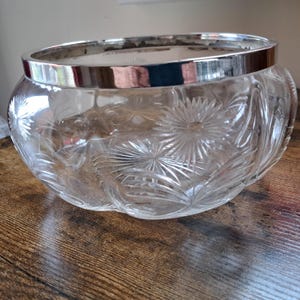 May include: A clear glass bowl with a silver rim. The bowl features a detailed, starburst pattern etched into the glass. The round bowl sits on a wooden surface.