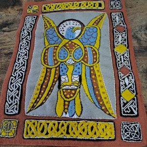 Może przedstawiać: Ręcznie szyta wisząca ozdoba ścienna z filcu z geometrycznym wzorem niebieskiego i żółtego ptaka. Ptak otoczony jest ozdobną ramką w kolorze żółtym i czarnym.