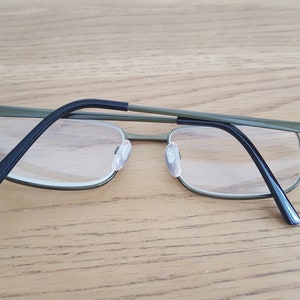 Op de afbeelding: Een bril van groen metaal met zwarte pootjes. De bril heeft heldere glazen en ligt op een houten oppervlak.