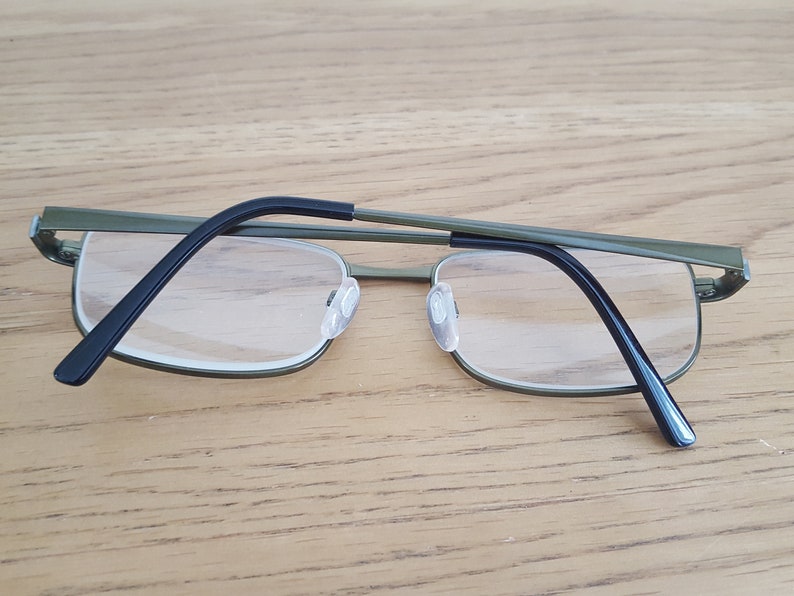 Op de afbeelding: Een bril van groen metaal met zwarte pootjes. De bril heeft heldere glazen en ligt op een houten oppervlak.