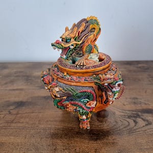 May include: A round, orange-toned incense burner featuring intricate, multi-colored dragon motifs in green, red, and gold. The lid is topped with a sculpted dragon figure, and the base is supported by three legs, all decorated with detailed relief patterns.