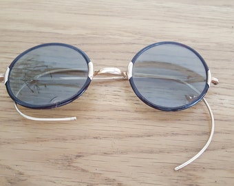 Könnte beinhalten: Eine runde Brille mit schwarzem Rahmen und goldenen Metallbügeln. Die Brille hat einen Vintage-Stil und liegt auf einer Holzoberfläche.