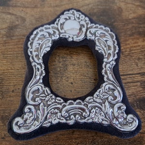 May include: A silver picture frame with an ornate design. The frame is on a dark brown surface.