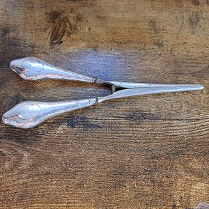 May include: Silver-toned serving tongs with a hammered texture on the handles and a spring mechanism. The tongs are designed with a curved shape and pointed ends, likely for serving food.