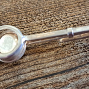 May include: A silver-toned metal tie clip featuring a circular setting at one end. The clip has a smooth, polished finish and a long, slender bar design that extends from the circular head, providing a secure grip for fastening onto a necktie.