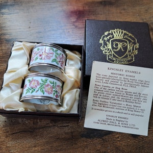 May include: Two silver napkin rings with white porcelain and a floral design of pink roses and green leaves. The rings are in a box with a label that reads "Kingsley Enamels Historic Roses Made in England by Alfred Hall & Sons Ltd. Stoke Prior, Bromsgrove, Worcestershire."