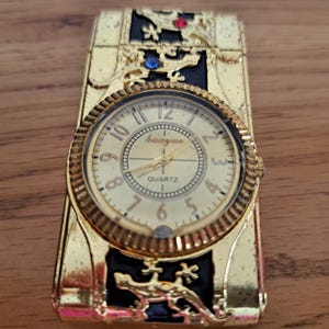 May include: Gold-toned quartz watch with a rectangular shape. The watch features a round face with gold numerals and hands, and is adorned with lizard-like designs and jewel accents. The watch has the word "Quartz" on the face.
