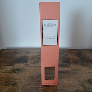 May include: A tall, rectangular, peach-colored box for a Pecksniff's England fragrance diffuser. The label reads Est. 1983 PECKSNIFF'S ENGLAND GINGER FLOWER & PATCHOULI FRAGRANCE DIFFUSER. A small cutout at the bottom reveals the glass bottle inside.