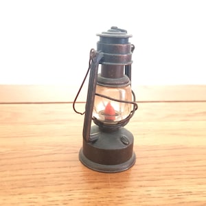 May include: A miniature antique lantern with a clear glass globe and a red light inside. The lantern is made of dark metal and has a handle for carrying. The lantern is a decorative item.