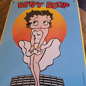 May include: A vintage-style metal sign featuring Betty Boop in a white dress with a red heart on the bodice. The background is a blue sky with a large orange sun behind Betty Boop. The sign reads "BETTY BOOP" in black letters.
