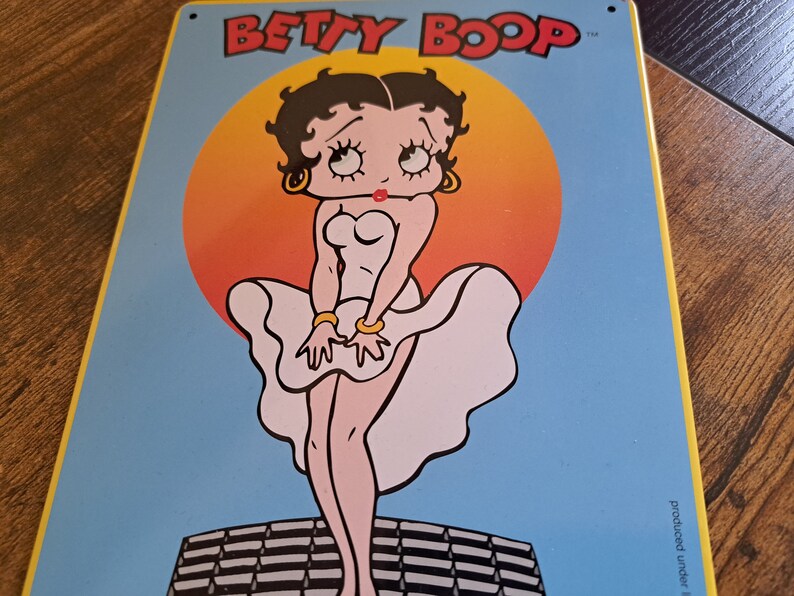 May include: A vintage-style metal sign featuring Betty Boop in a white dress with a red heart on the bodice. The background is a blue sky with a large orange sun behind Betty Boop. The sign reads "BETTY BOOP" in black letters.