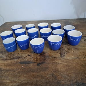 Puede incluir: Un conjunto de tazas de cerámica azules con interiores blancos. Cada taza tiene un nombre de lugar diferente escrito en escritura blanca. Las tazas están dispuestas sobre una superficie de madera oscura. Las tazas miden aproximadamente 6 cm de alto.