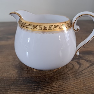 May include: White ceramic creamer with a gold and black geometric design around the rim.