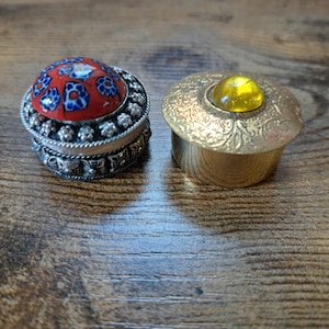 May include: Two small decorative boxes. One has a red and blue floral design on the lid and a silver-colored base. The other is gold-colored with a yellow gem on top. Both are on a wooden surface.