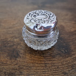 May include: A small, clear glass jar with a silver lid. The lid has a decorative design of swirling leaves and flowers.