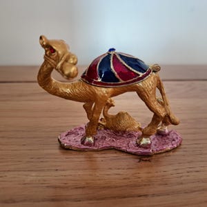 Op de afbeelding: Een goudkleurig kameelfiguur met een decoratief zadel in rood, blauw en goud. De kameel staat op een roze basis met een kleinere kameelfiguur. Een kleine blauwe edelsteen bevindt zich bovenop het zadel.