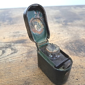 Op de afbeelding: Een open, antieke inktpot met een scharnierend deksel. De inktpot is zwart met een groen interieur en een kleine, ronde inktpot. Het deksel heeft een decoratief element met tekenen van ouderdom en slijtage. De inktpot staat op een houten oppervlak.