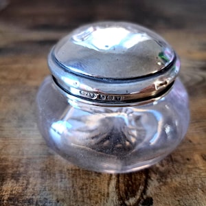 Può includere: Un piccolo barattolo di vetro trasparente con un corpo arrotondato e un coperchio argentato. Il coperchio è a cupola e si adatta perfettamente al barattolo. Il barattolo sembra essere antico, con un'estetica vintage. Il barattolo è su una superficie di legno.