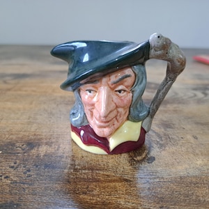 May include: A ceramic pitcher with a detailed face of an elderly man wearing a dark green hat. The handle is shaped like a walking stick. The man's face is painted with realistic features, including wrinkles and a long nose. The pitcher has a burgundy and yellow collar.
