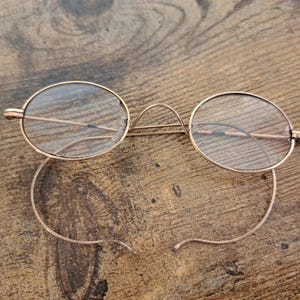 May include: Antique gold-tone eyeglasses with oval lenses. The glasses feature a delicate bridge and wire temples, resting on a wooden surface. The lenses show some wear, adding to the vintage aesthetic. A classic eyewear accessory.