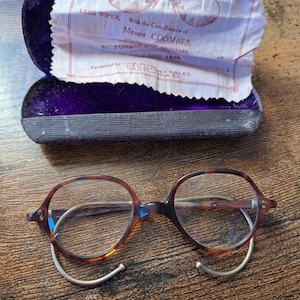 Könnte beinhalten: Eine Brille aus Schildpatt mit runden Gläsern und silbernen Bügeln. Die Brille liegt auf einer Holzoberfläche. Die Brille wurde mit einem Linsenwischer von Messrs. Coombes, Optiker, 87, Tottenham Court Road, London, 1808, geliefert.