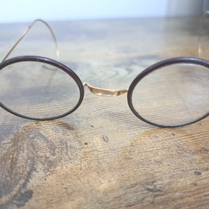 May include: A pair of vintage round spectacles with a dark brown frame and gold-coloured metal arms. The clear lenses sit on a wooden surface. The design is reminiscent of antique eyewear.