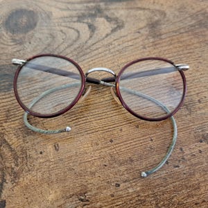 May include: Round eyeglasses with burgundy frames and silver-colored metal accents. The lenses are clear, and the temples are a light green color. The glasses are resting on a wooden surface.