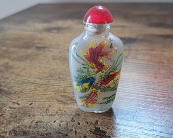 May include: A small, clear glass bottle with a red stopper. The bottle is decorated with colorful images of goldfish and aquatic plants. The bottle is likely used for perfume or essential oils.