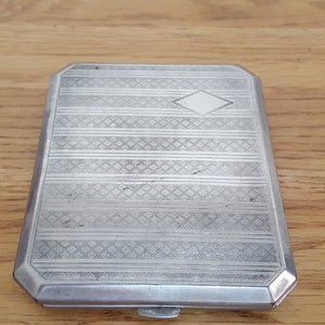May include: A silver cigarette case with a diamond-shaped design in the center and a patterned design on the lid.