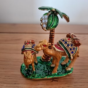 Op de afbeelding: Een decoratief beeldje met twee kamelen en een palmboom. De kamelen zijn bruin met juweelkleurige zadels. De palmboom heeft groene bladeren en een cluster van witte parels. De basis is groen en het beeldje heeft gouden accenten.