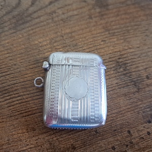 May include: A silver match safe with an ornate engraved design. The match safe has a hinged lid and a small loop for carrying.