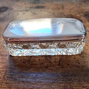 Può includere: Una scatola rettangolare antica placcata in argento con base in cristallo tagliato. Il coperchio è inciso con un motivo floreale e la base presenta un motivo a taglio diamante. La scatola viene probabilmente utilizzata per riporre piccoli oggetti.
