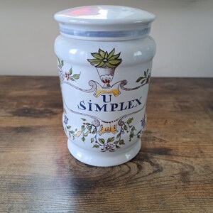 May include: White ceramic jar with a blue and white lid. The jar features a floral design in yellow, green, and blue. The jar is labelled "U SIMPLEX".