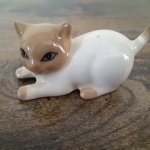 May include: A white porcelain figurine of a cat with brown markings. The cat is lying on its belly with its head turned to the side. The cat has black eyes and a brown nose.