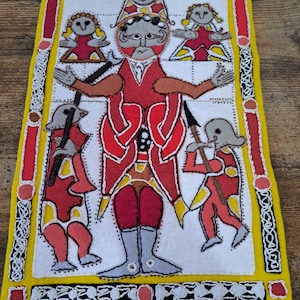 May include: A white felt tapestry with a red, yellow, and black border. The tapestry features a central figure with a red robe and a crown, surrounded by smaller figures. The figures are embroidered in a simple style with white thread.