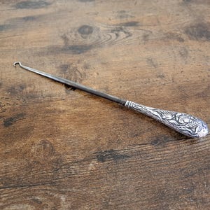 May include: A vintage crochet hook with a silver-toned handle featuring an ornate floral design. The hook is made of dark metal and is lying on a wooden surface. The hook is a tool used for creating crochet projects.