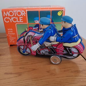 Könnte beinhalten: Vintage Spielzeugmotorrad mit zwei Fahrern in blauen Uniformen. Das rot-rosa Motorrad hat ein Seitenrad zum Ausbalancieren. Auf der Box steht "MOTOR-CYCLE" und "With siren Friction For children ages 3-up."