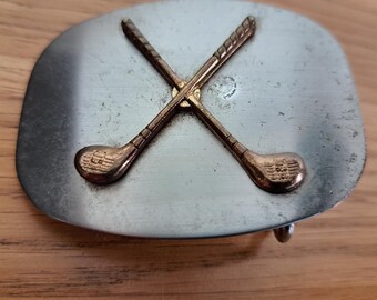 May include: A silver-toned metal belt buckle with two crossed golf clubs in a gold color. The buckle has a rectangular shape with rounded edges and a hook on the back for attaching to a belt. The golf clubs are detailed with textured grips and club heads.