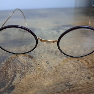 Op de afbeelding: Een ronde bril met een gouden montuur en donkerbruine pootjes. De bril ligt op een houten oppervlak.