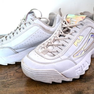 以下が含まれることがあります： 白のFila Disruptor IIスニーカーのペア。分厚いソール、レースアップ開閉、黄色のFilaロゴが特徴です。スニーカーは真珠色のアクセントがあり、木製の表面に置かれています。