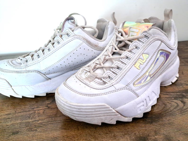 以下が含まれることがあります： 白のFila Disruptor IIスニーカーのペア。分厚いソール、レースアップ開閉、黄色のFilaロゴが特徴です。スニーカーは真珠色のアクセントがあり、木製の表面に置かれています。