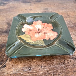 May include: A square, dark olive green ceramic ashtray featuring a central painted illustration of a large, light orange hibiscus flower with green leaves. The ashtray has four notched corners and shows minor blue paint wear on the bottom edge.