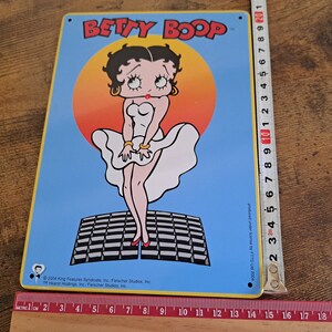 May include: A blue and yellow metal sign featuring Betty Boop in a white dress, standing on a black and white checkered platform. The sign reads "BETTY BOOP" in yellow letters.