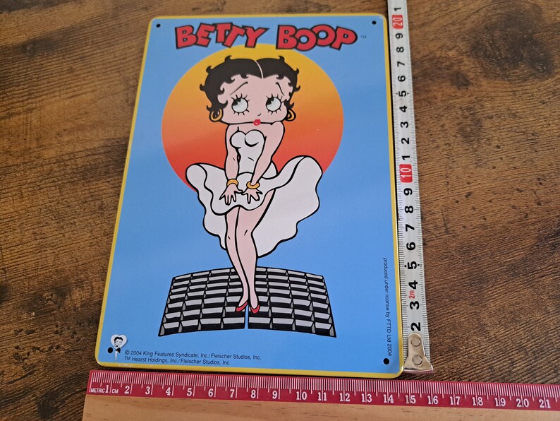 May include: A blue and yellow metal sign featuring Betty Boop in a white dress, standing on a black and white checkered platform. The sign reads "BETTY BOOP" in yellow letters.